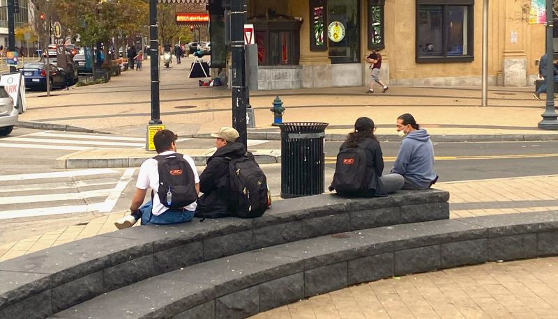 Four people, two with backpacks on, sit on a concrete bench in the Columbia Heights civic plaza. Their backs are turned to us and each pair are in conversation.