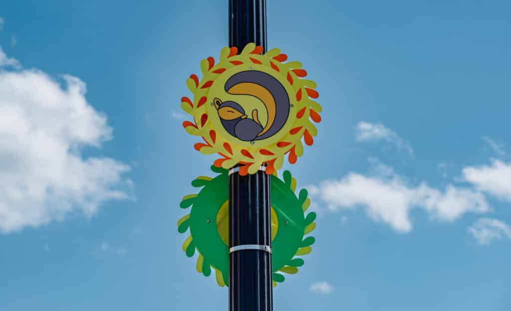 A black metal pole has a green and orange sculpture of leaves in a circle with a grey and brown squirrel centered in the middle with its tail wrapped around it. The sky is blue with white puffy clouds behind it. 