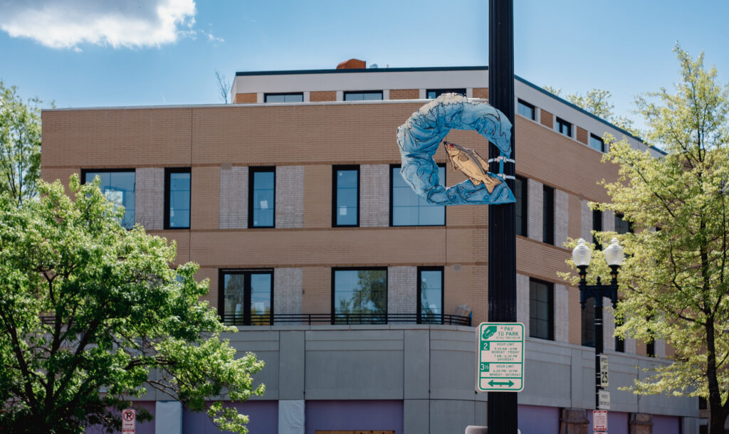 a large pol with parking signs on it displays a blue sculpture of water with a fish leaping out of the center. Behind the pole is a beige office building. The sky is blue and there are white puffy clouds in the distance.