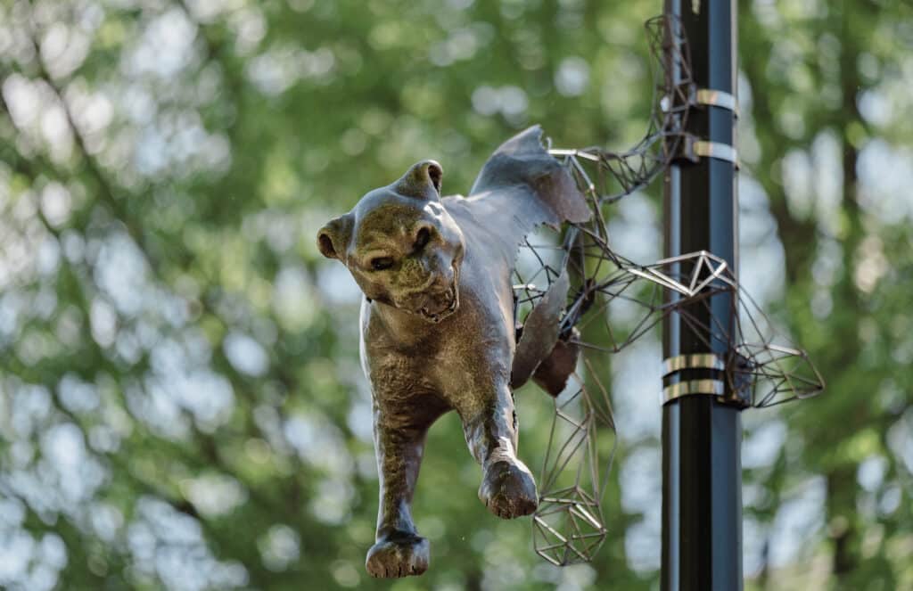 A sculpture of a large cat with an aggressive growling face is leaping from a pole. We see trees leaves in the background.