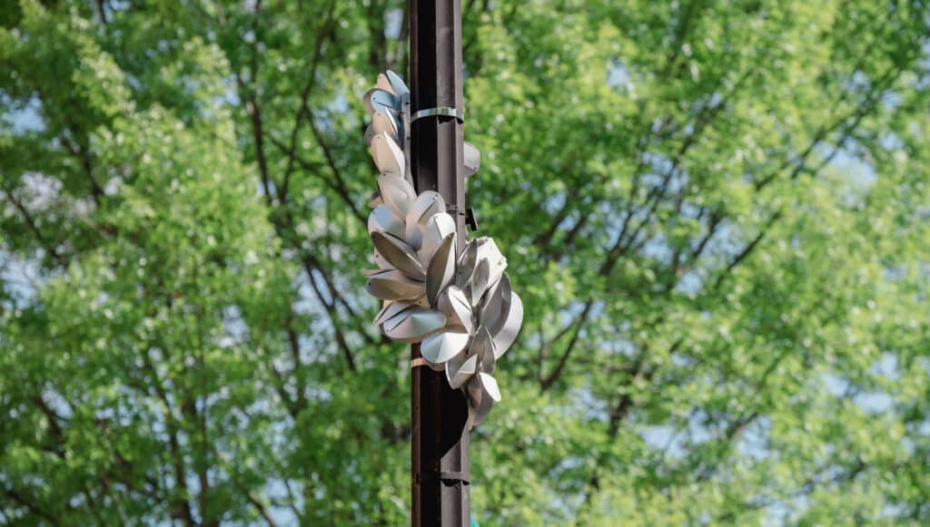 a metal pole with a silvery-metal sculpture of things that look like barnacles crawling up the pole. There is a large green tree in the background.